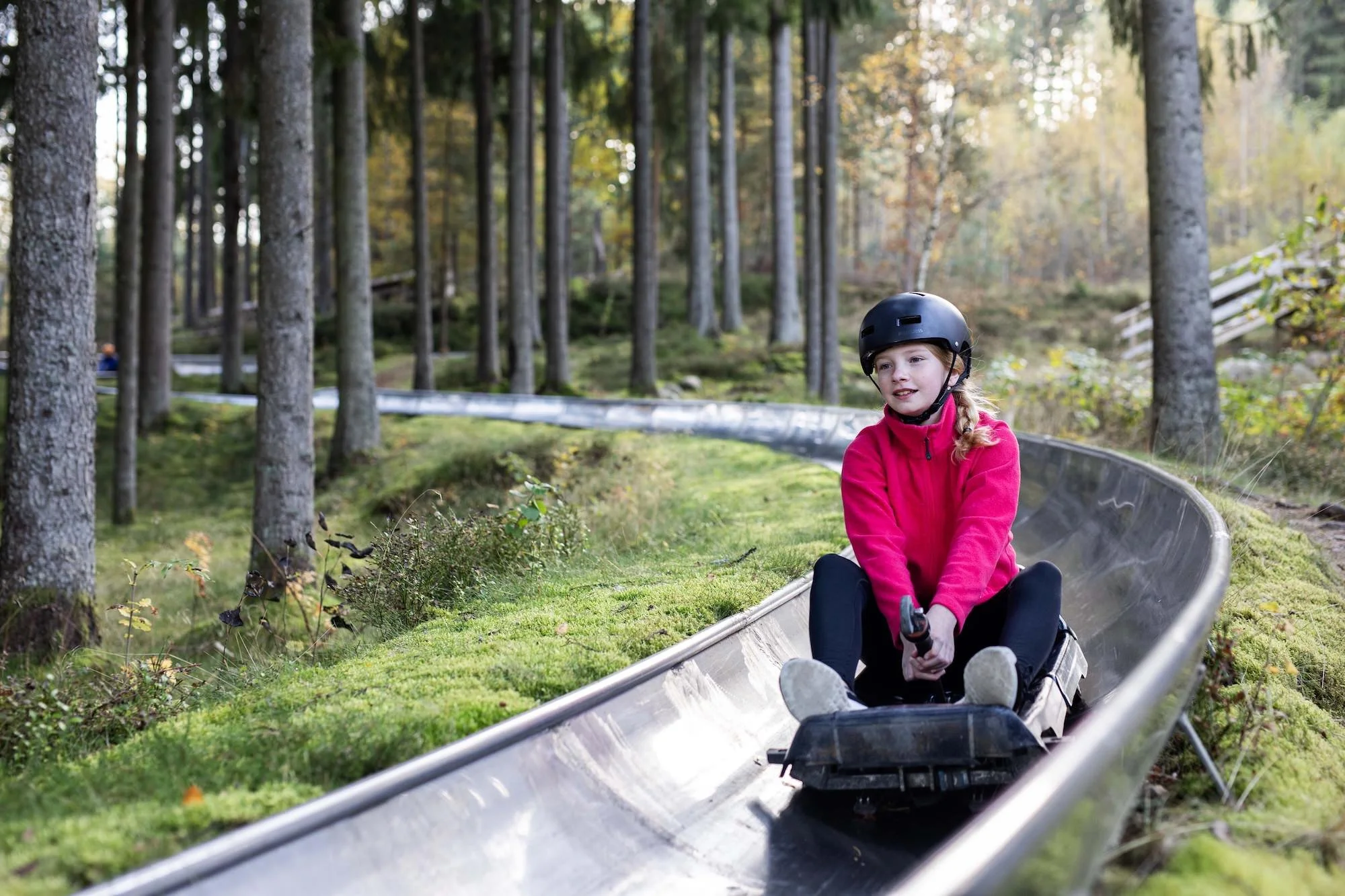 En flicka i en röd jacka och svart hjälm åker i en rodelbana i skogen på hösten.