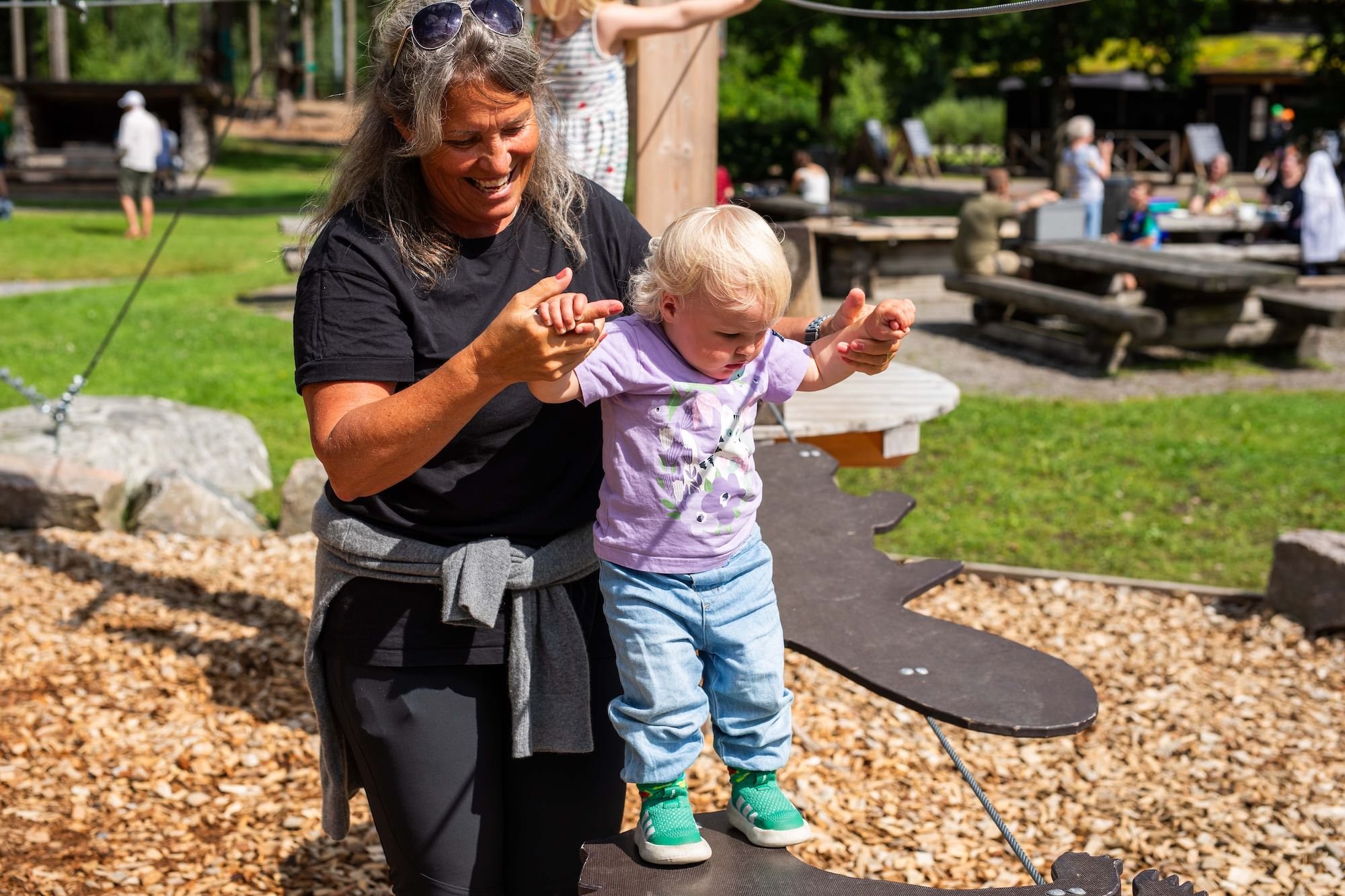 En kvinna och ett litet barn leker på en lekplats på en solig dag, med grönt gräs och andra barn i bakgrunden.