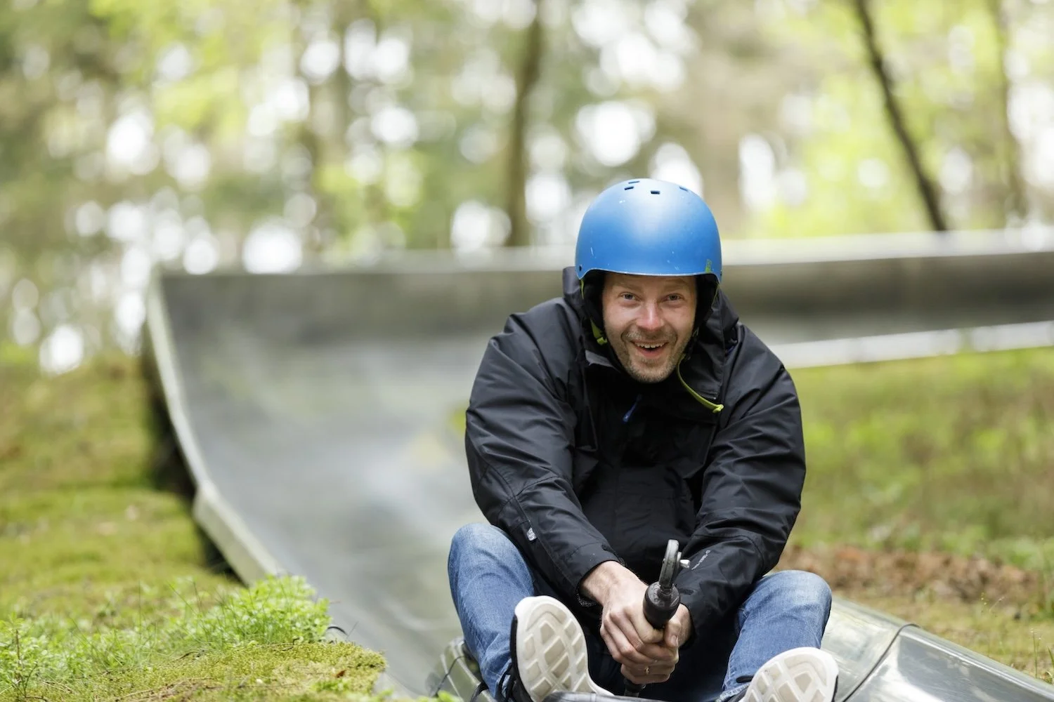 Man med blå hjälm som åker på en plastskidlift i skogsområde, ler och håller i ett handtag.