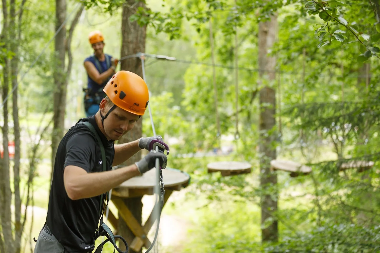 Två personer som utför repklättring i skogen, båda med orange hjälmar och säkra utrustningar.