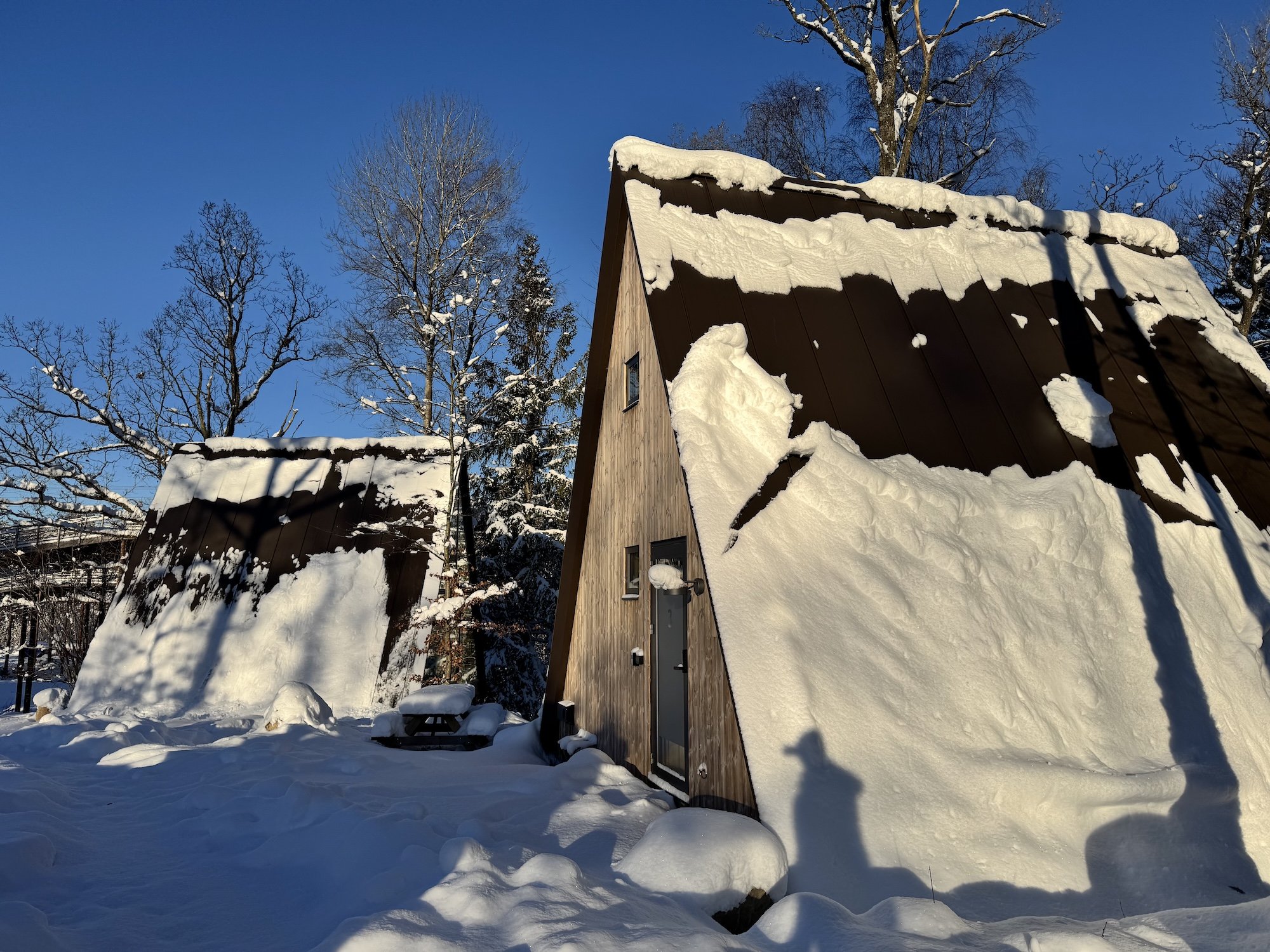 Två stugor med snötäckta tak, en större till höger och en mindre till vänster, under en klar blå himmel. Träd utan löv i bakgrunden och snö på marken.