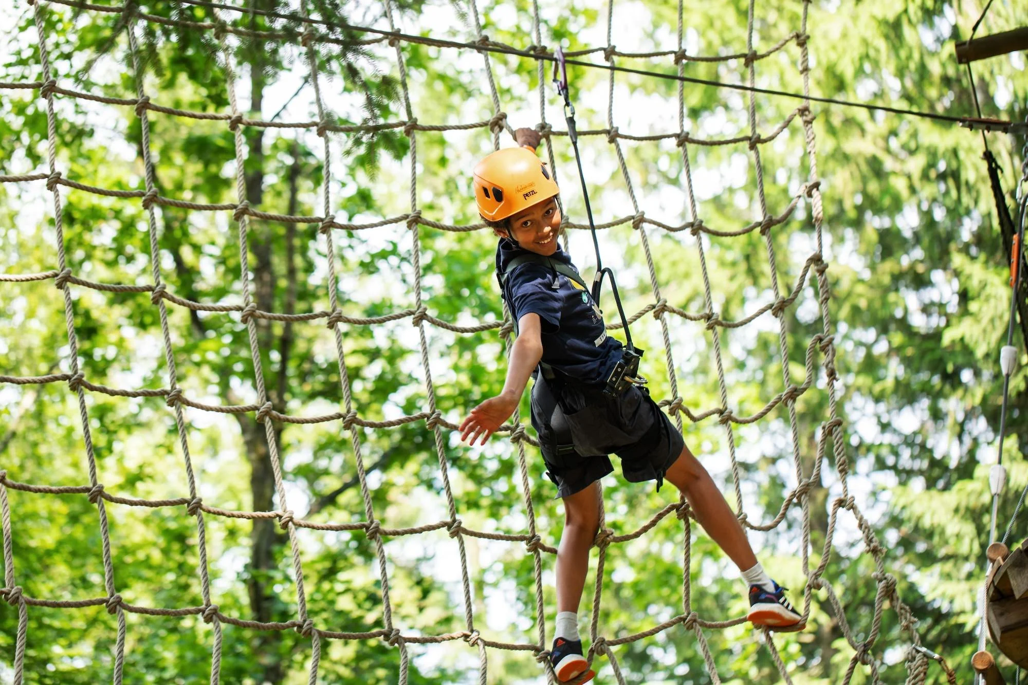Ung pojke i hjälm och säkerhetsutrustning går på ett grovt nät i ett trädtoppsträd. Blad och träd i bakgrunden.