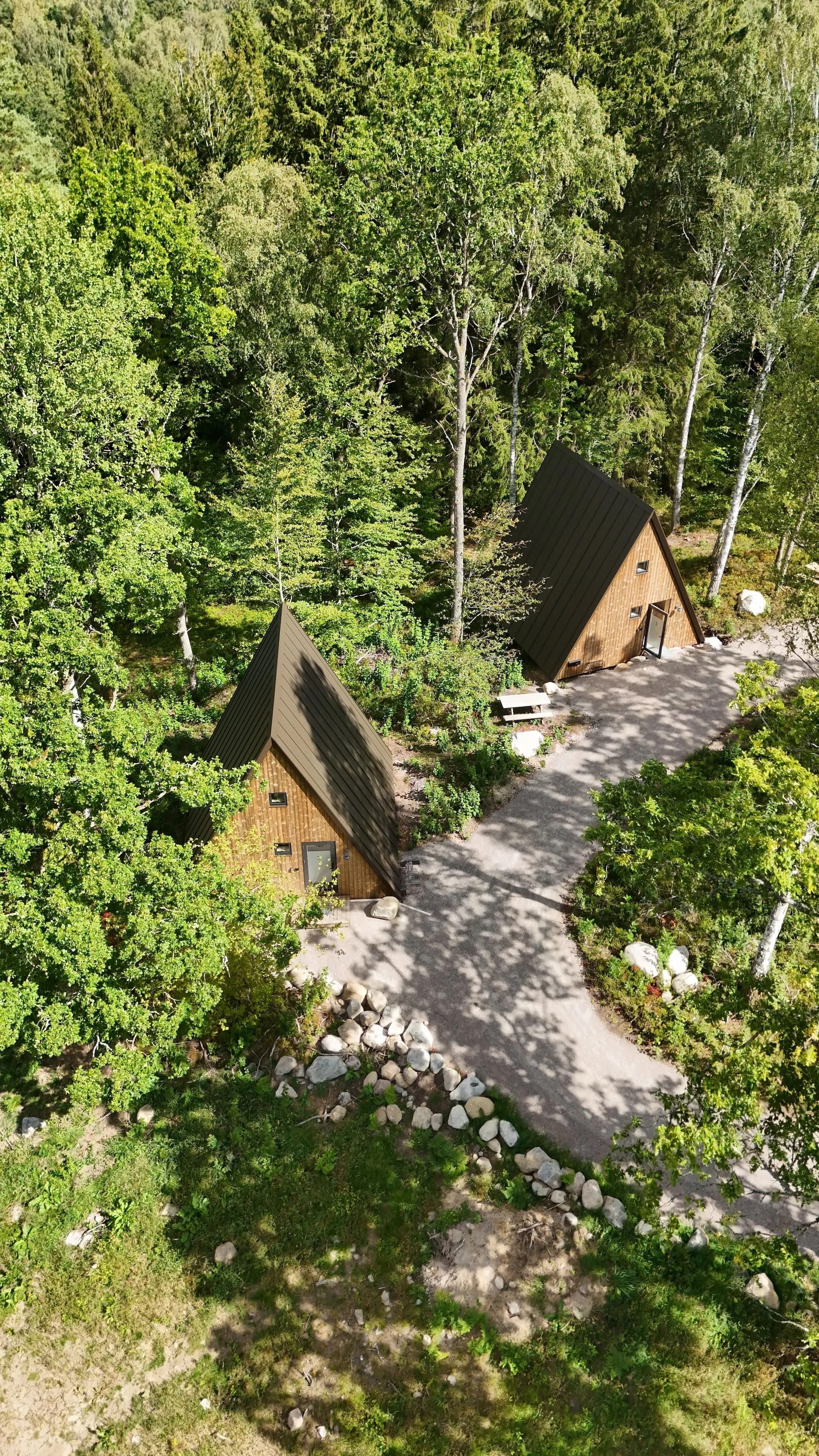 Två A-frame-hus med branta tak, omgivna av gröna träd, en grusgång och stenar i en skogsmiljö.