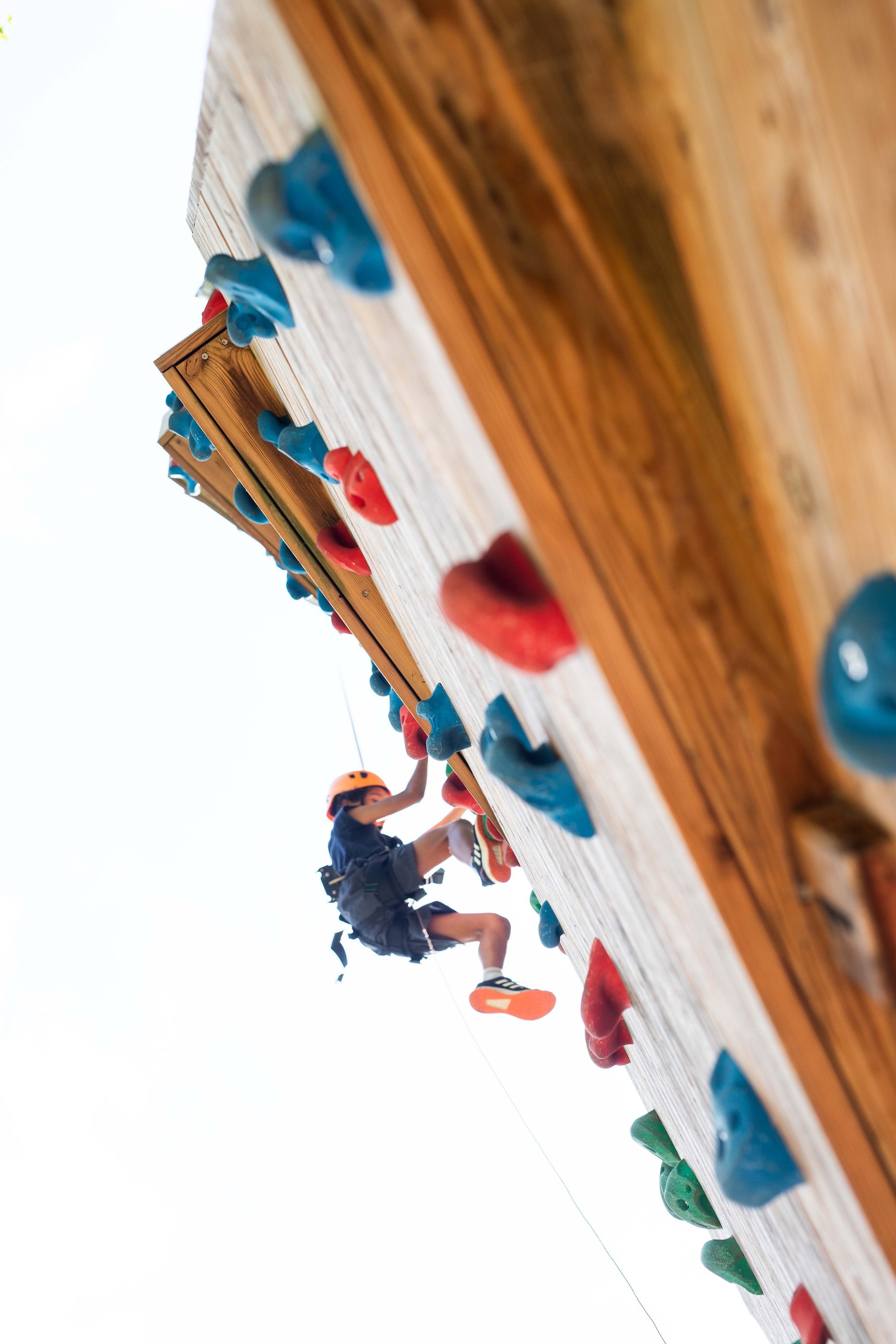 Person som klättrar uppför en utomhusklättervägg med färgglada hand- och fotgrepp, iklädd hjälm och säkerhetssele.