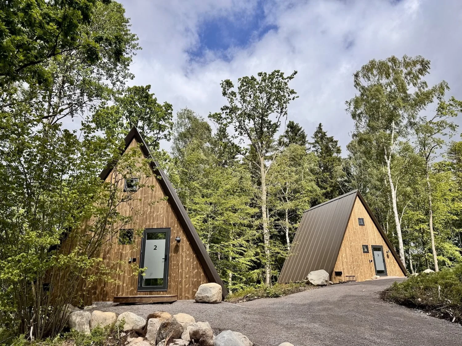 Två moderna A-formade stugor med träfasad, omgivna av träd och bergets naturliga landskap.