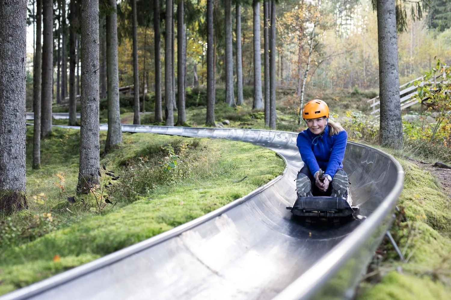 Ung kvinna i säkerhetsutrustning åker alpinbedåbana i en skog, klädd i blå jacka och orange hjälm.