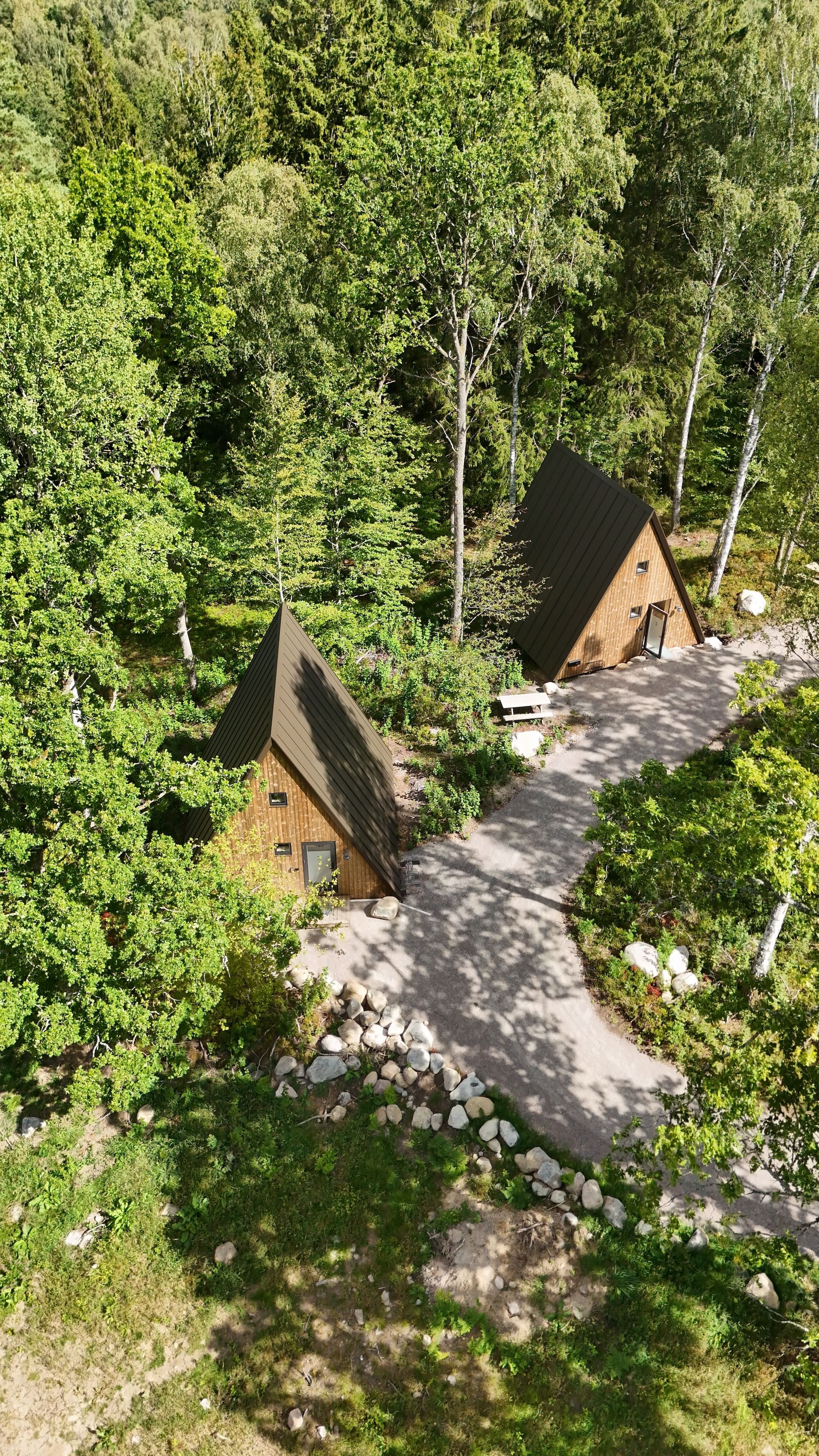 Två små trähus med branta tak ligger i en lövskog, med en grusad gångväg och omgivande stenbumlingar.