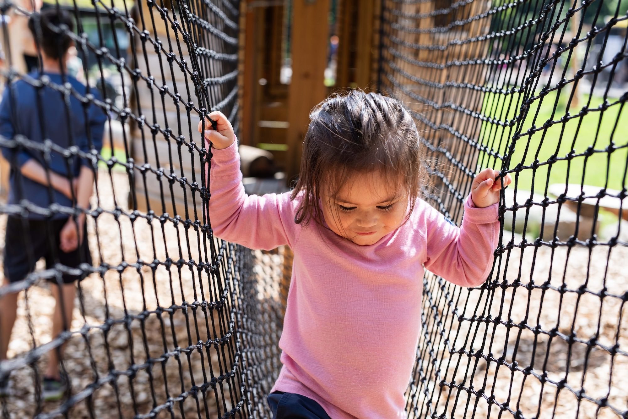 En liten flicka i rosa tröja som kryper genom ett nät på en lekplats medan en pojke i bakgrunden tittar bort.
