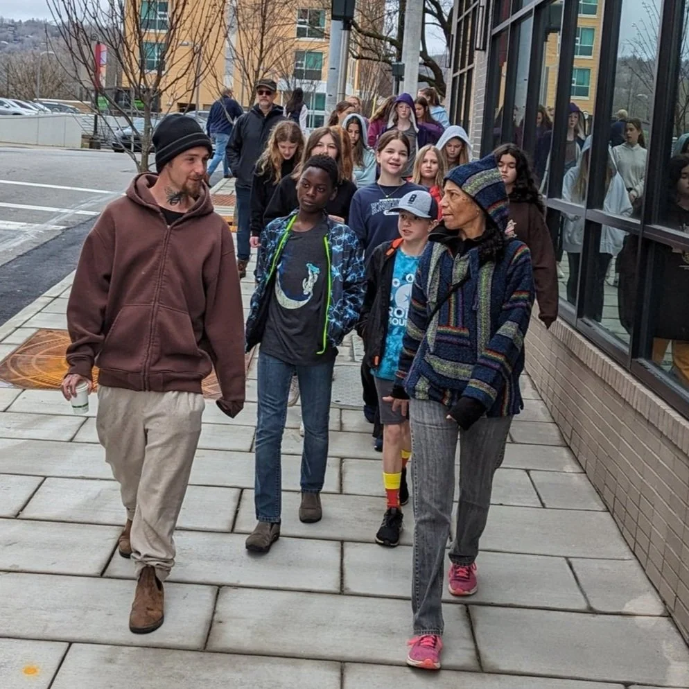 Group of people walking on the sidewalk, including a man, a woman, children, and teenagers, near a large glass window in an urban area during the daytime.