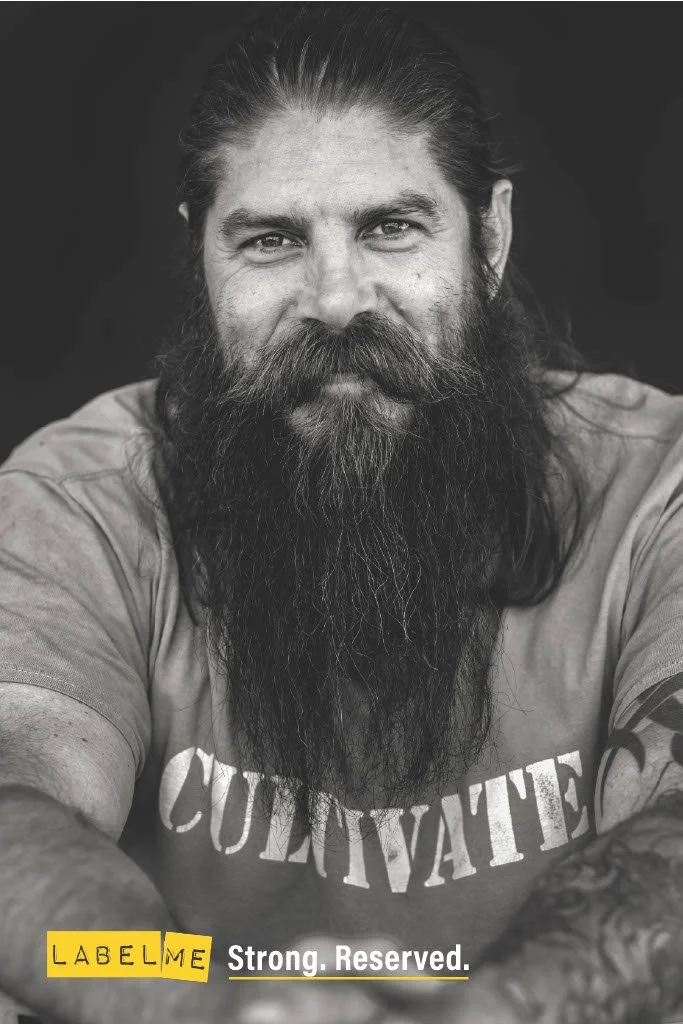 Black and white portrait of a man with long hair and a full beard, wearing a T-shirt with the word 'CONTENT' partially visible, and tattoos on his arms.