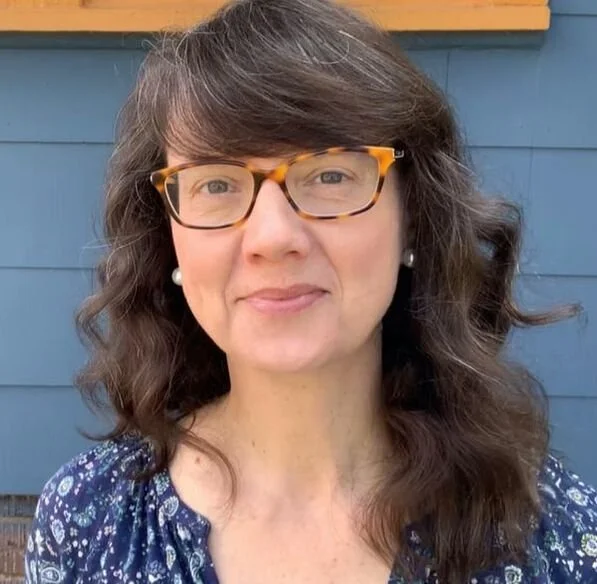 A woman with wavy brown hair, glasses with tortoiseshell frames, and pearl earrings, standing outside in front of a blue wall with a wooden trim.