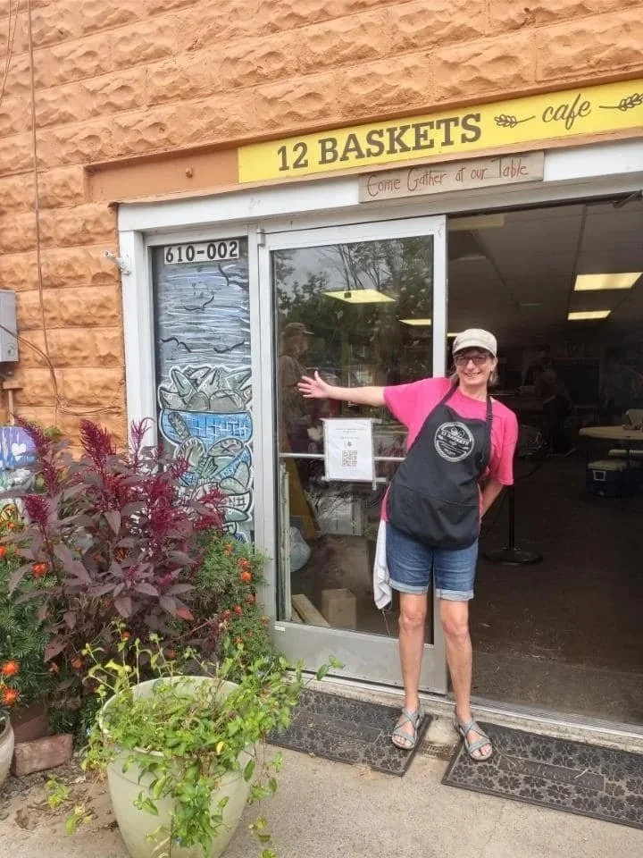 A woman smiling and gesturing towards a store entrance labeled '12 Baskets'. She is wearing a pink shirt, denim shorts, sandals, and a cap, with a black apron around her neck. The shop has a glass door with a reflection of trees and ceiling lights inside. There are potted plants and flowers outside the shop.