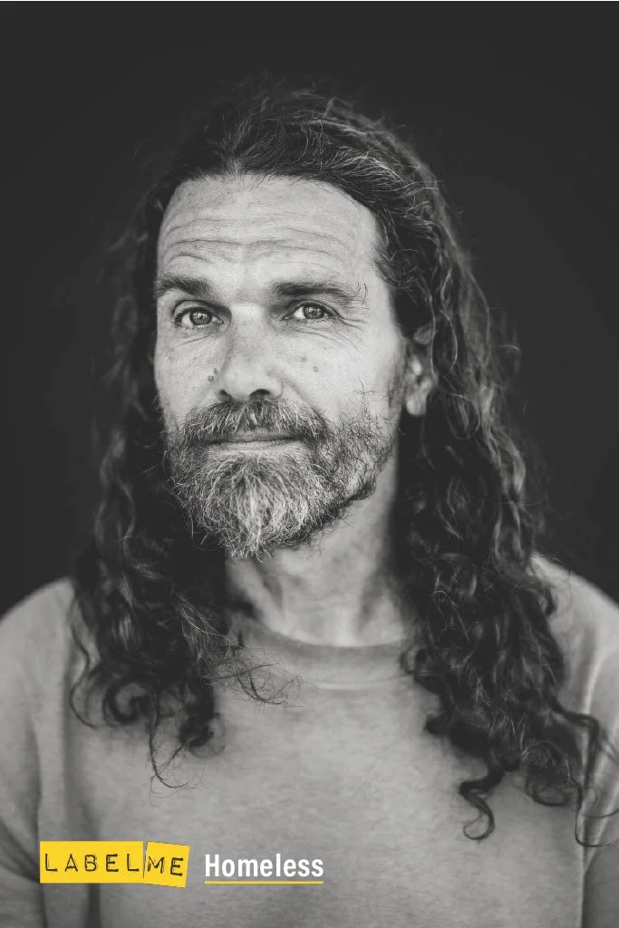 Black and white portrait of a middle-aged man with long curly hair, a beard, wearing a casual t-shirt, with text "LABEL ME Homeless" at the bottom.
