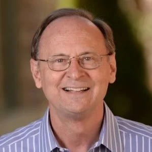 A smiling middle-aged man with glasses, wearing a blue striped shirt, standing outdoors.