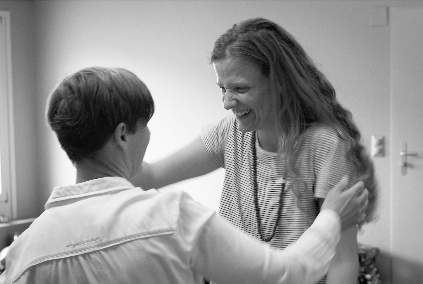 Two women smiling warmly at each other in a friendly embrace in a room with plain walls.