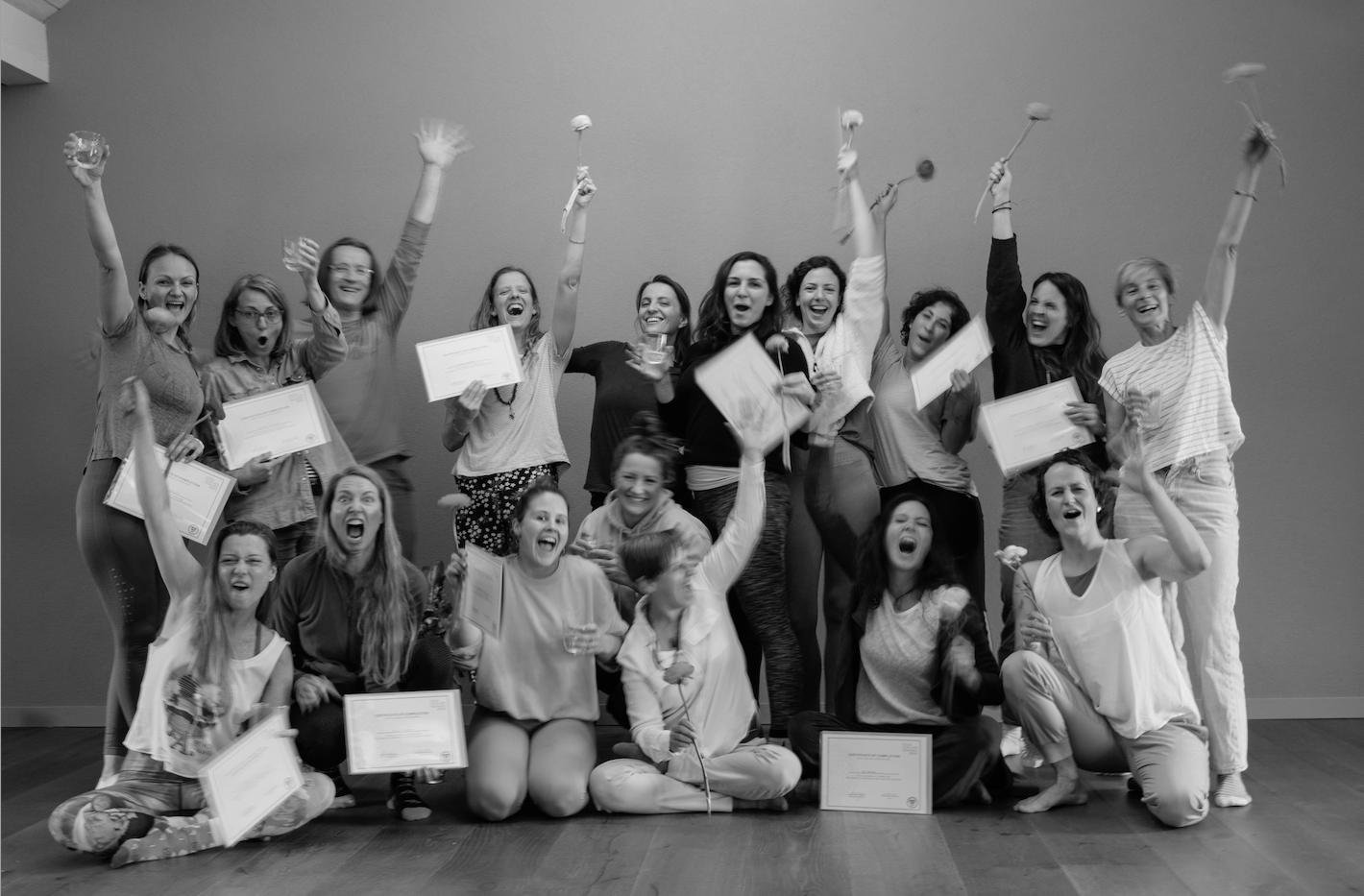 Black and white photo of a group of women celebrating with certificates and flowers, smiling and cheering, in a room with plain walls.