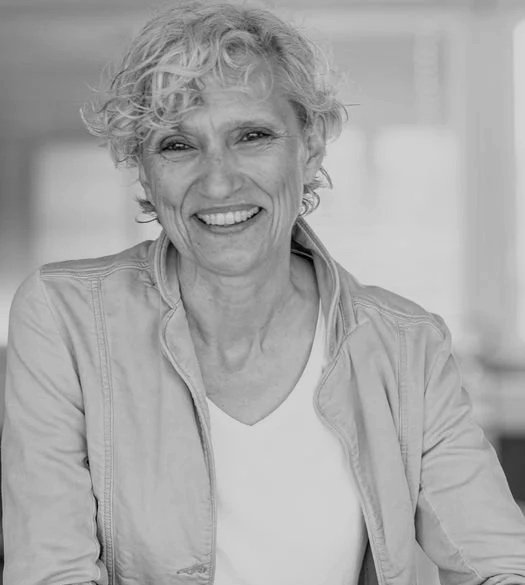 A smiling woman with short curly hair, wearing a light jacket over a white shirt, in a modern indoor setting.