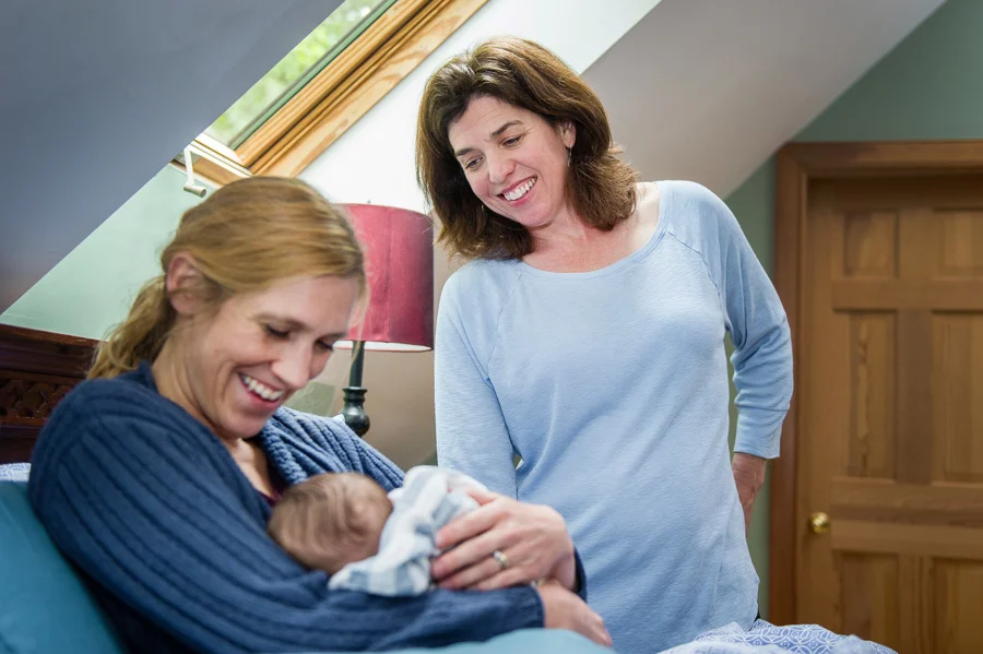 Two women smiling, one holding a newborn baby in a cozy room with natural light, wooden furniture, and a green wall.
