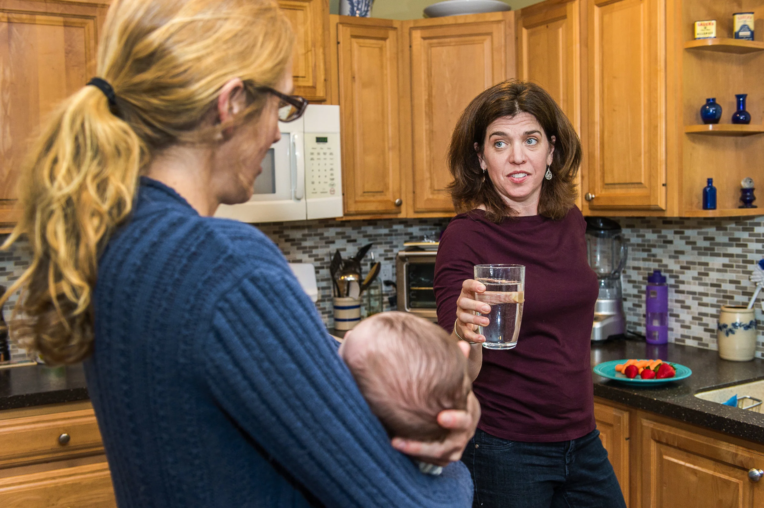 Two women and a baby in a kitchen, one woman with glasses and a ponytail, the other with shoulder-length brown hair holding a glass of water, ahead of a kitchen counter with a plate of strawberries and blueberries.
