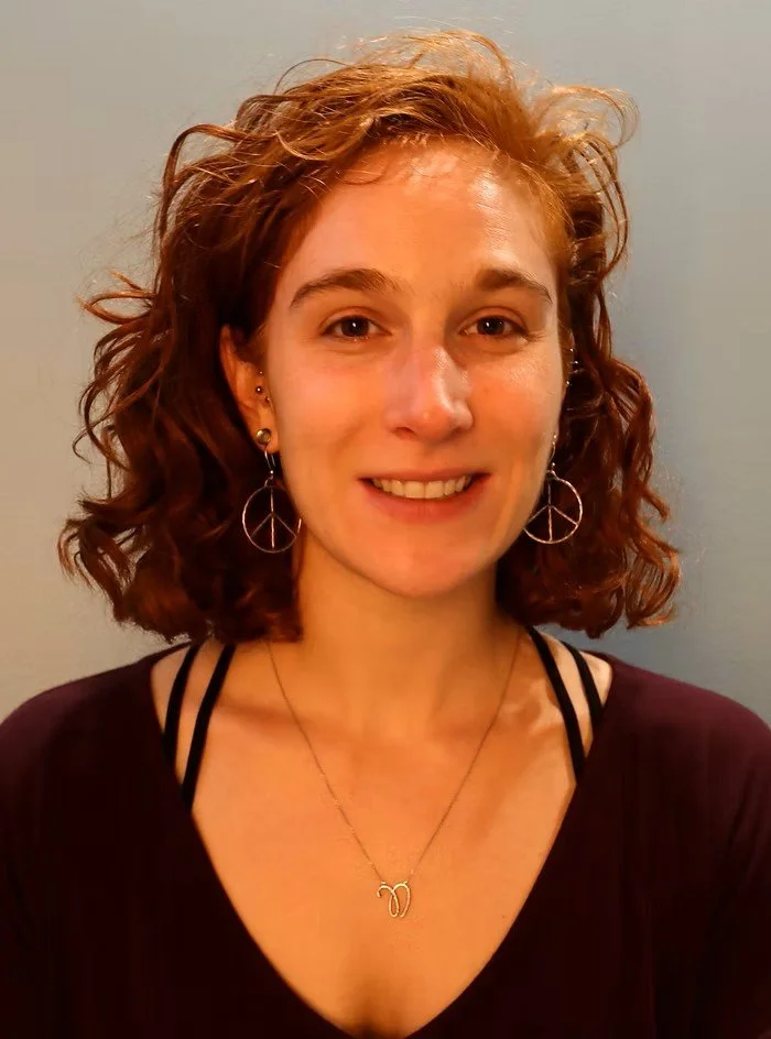 A young woman with curly red hair smiling at the camera, wearing peace sign earrings, a necklace with a double 'M' pendant, and a dark top with thin strap details.