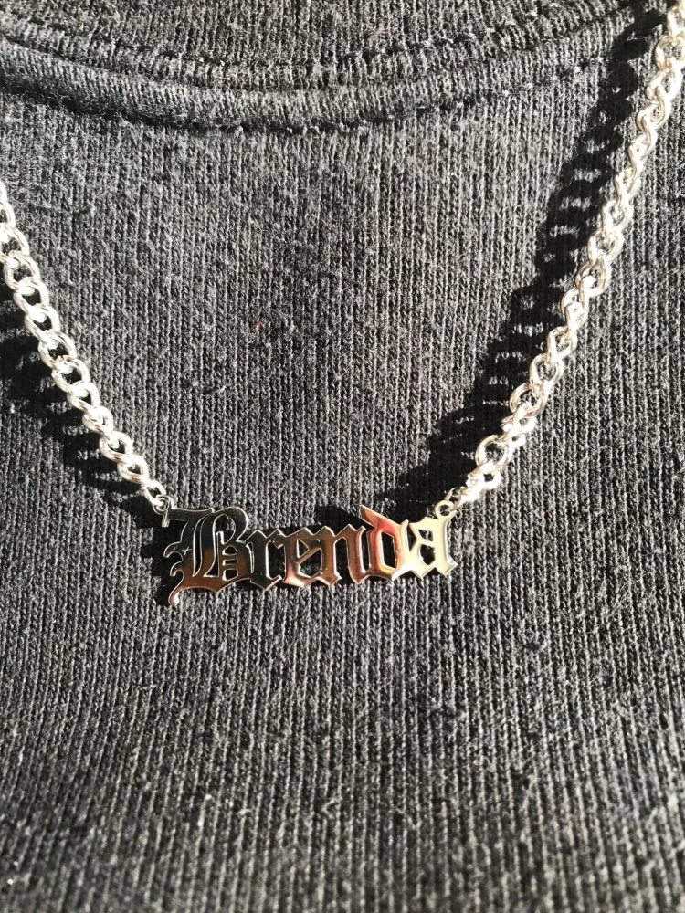 Silver chain necklace with the name "Brenda" in Gothic font on a black fabric background.