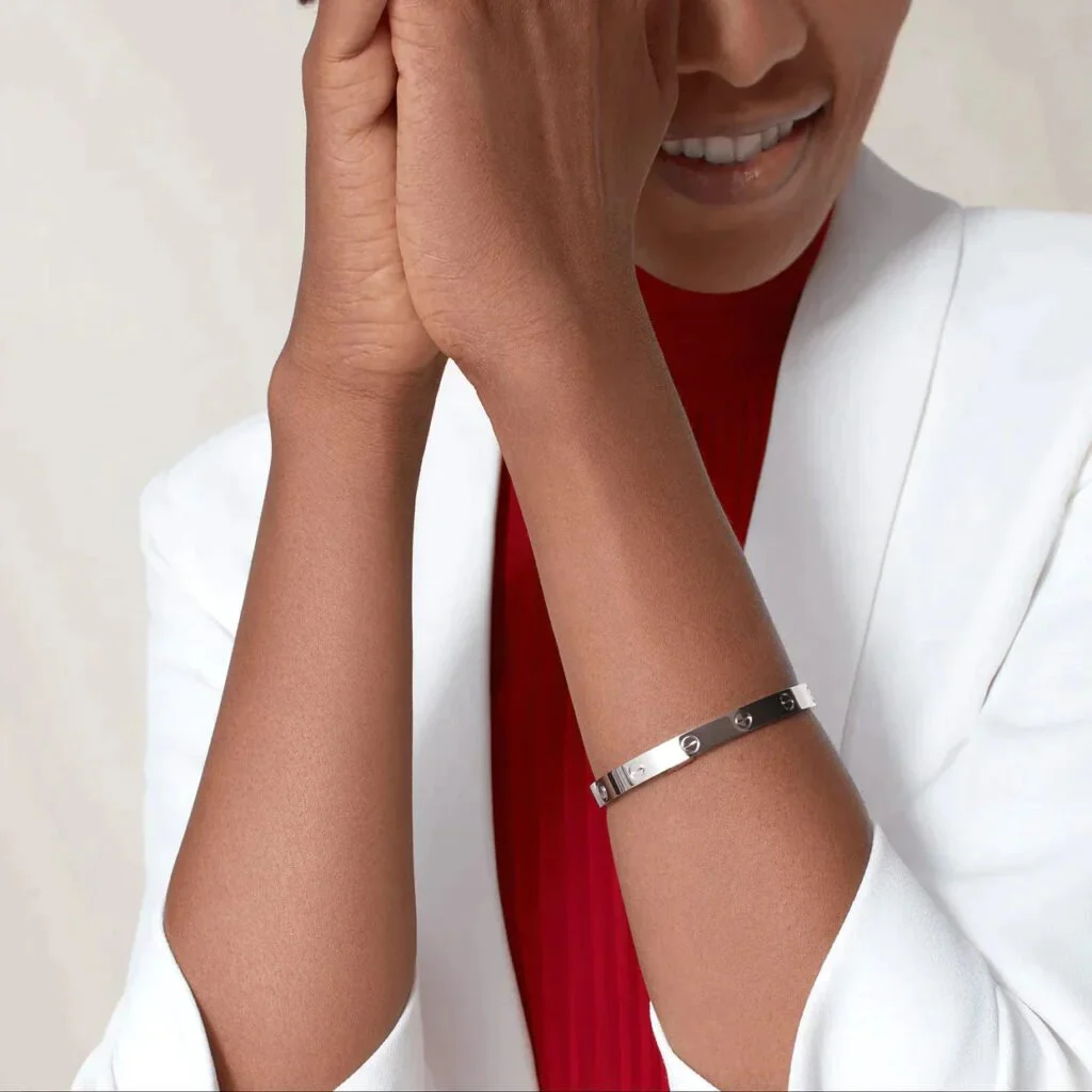 Person wearing a silver bracelet with screw motifs, dressed in a white jacket and a red top, covering their mouth with a hand.