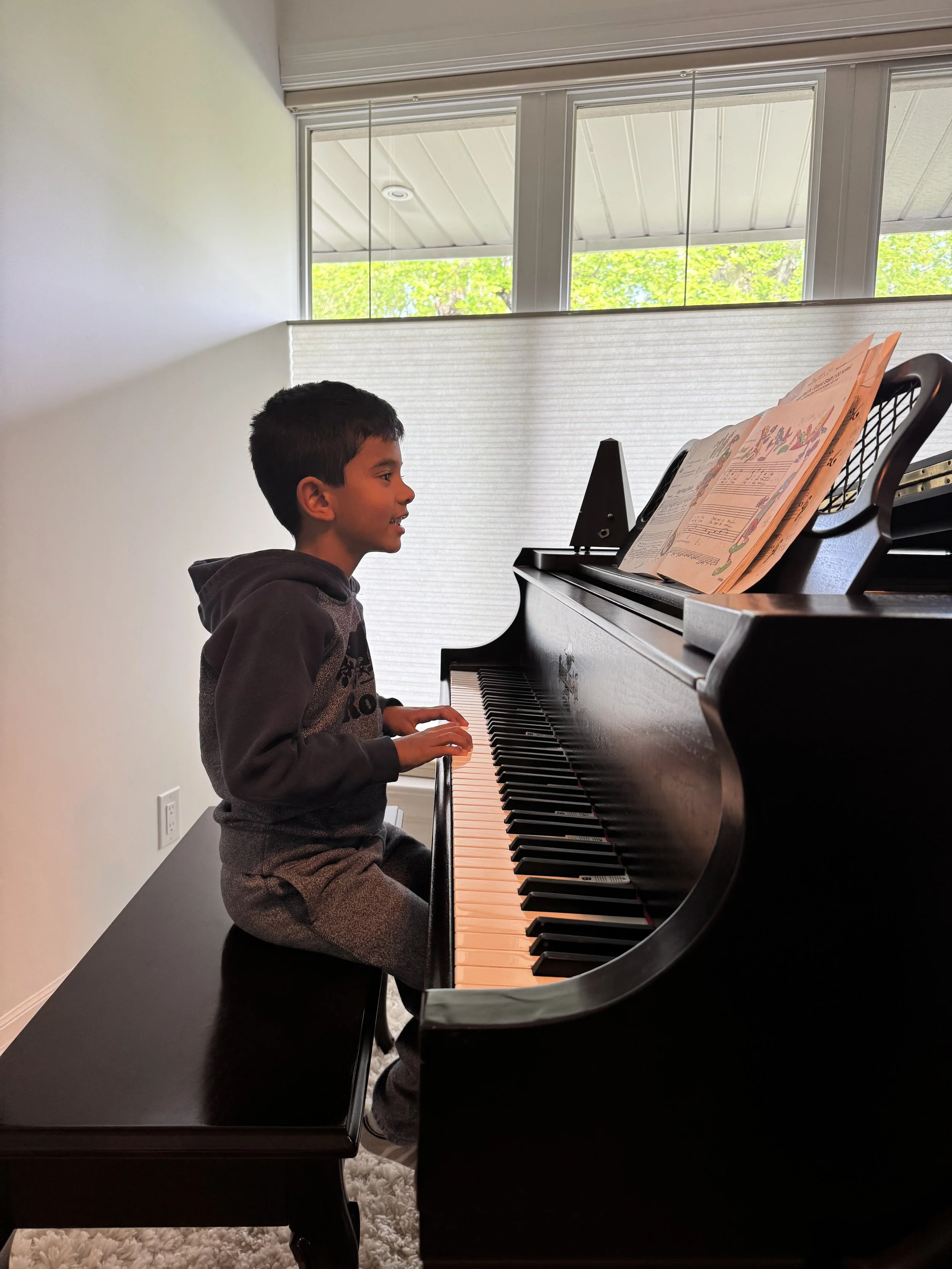A young boy sitting at a black grand piano, playing with sheet music on the music stand, in a room with large windows and white walls.