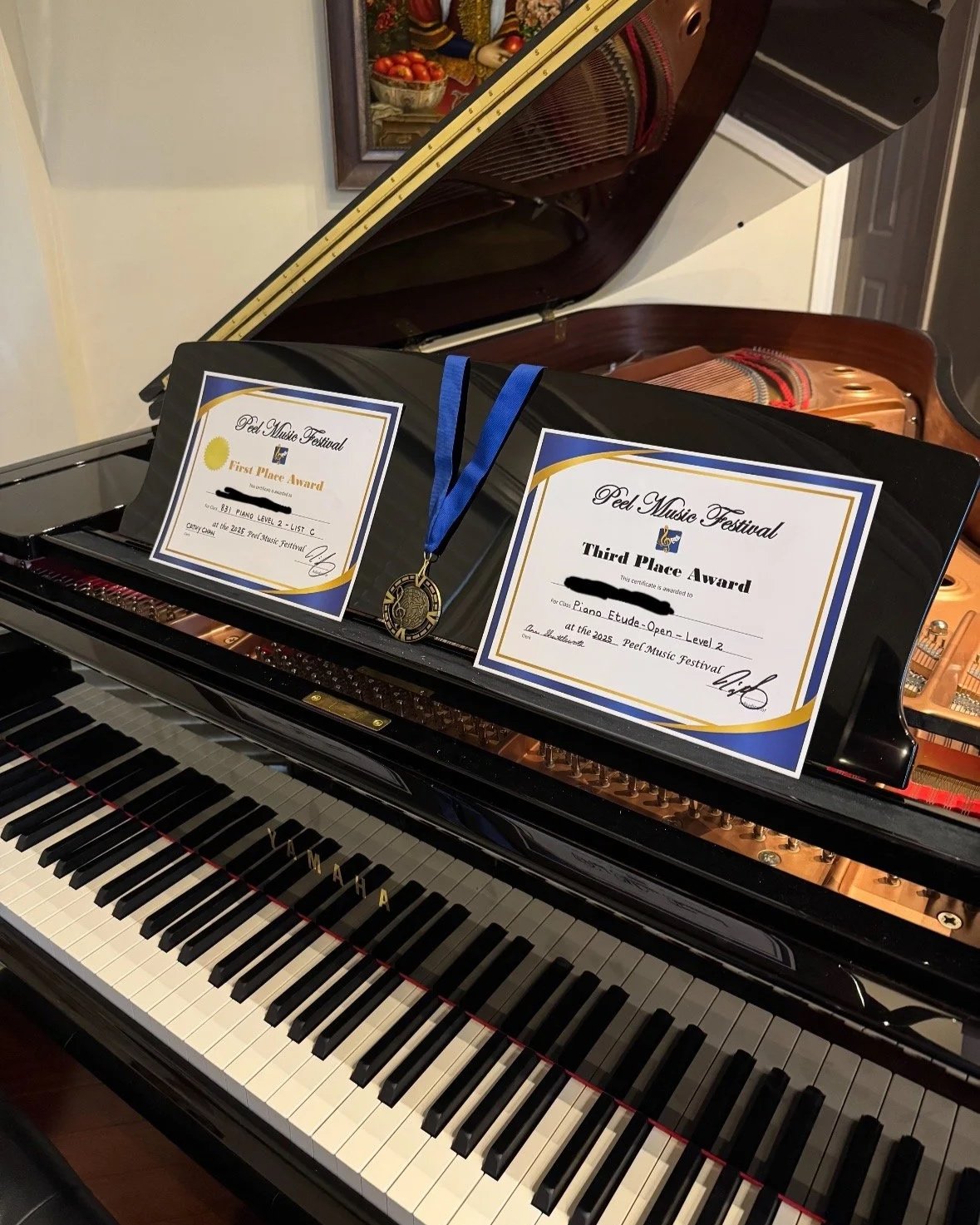 A grand piano with two award certificates and a medal on top. The certificates indicate third place and first place wins at the Peel Music Festival for piano competitions.