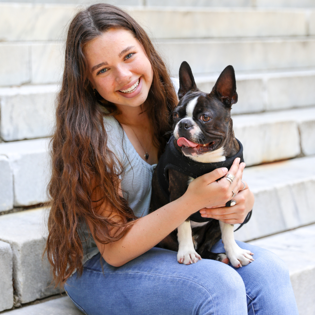 A young woman with long wavy hair and a bright smile sitting on outdoor steps, holding a black and white Boston Terrier dog wearing a harness, with her arm around the dog.