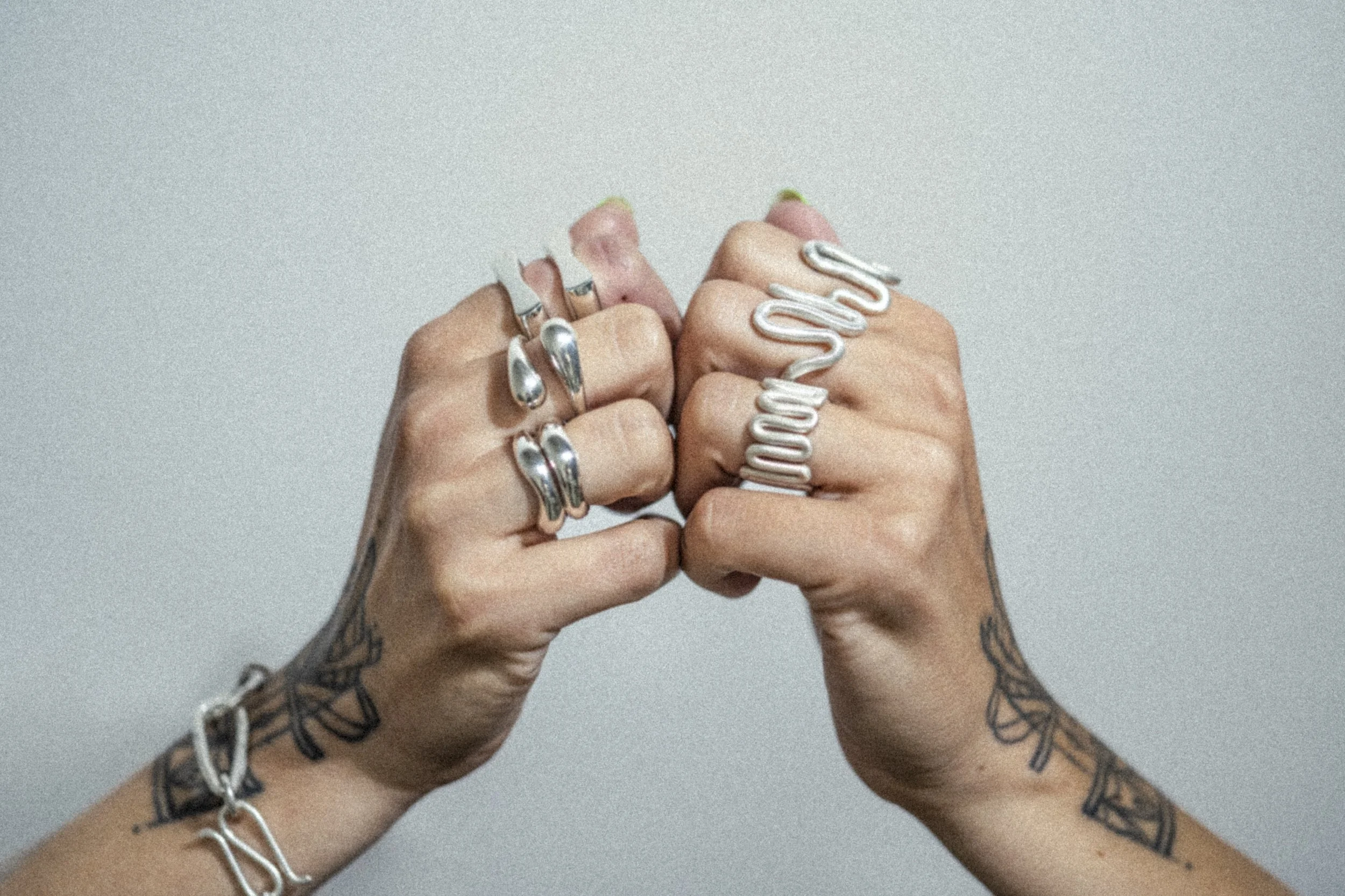 Zwei Hände mit silbernen Ringen und Tattoos in einem Handgelenk mit Schmuckstücken. Die Hände sind in Fäuste geballt, aufeinander gerichtet.