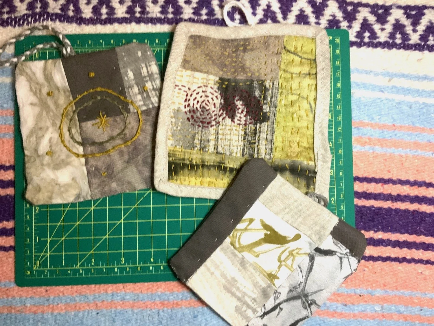 Three fabric pouches with abstract and artistic designs placed on a green cutting mat, which is on top of a multicolored striped carpet.