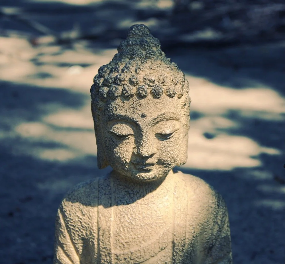 Sand sculpture of a Buddha with a peaceful expression, detailed features, and textured surface, placed outdoors on the sandy beach with shadows.