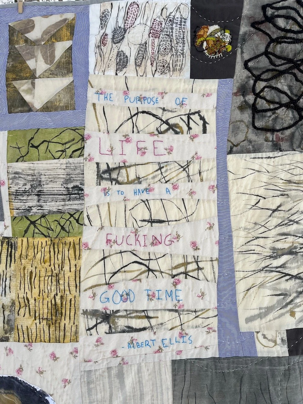 A quilt with various fabric pieces containing different abstract and nature-inspired patterns. In the center, there is embroidered text that reads: "The purpose of life is to have a fucking good time - Albert Ellis".