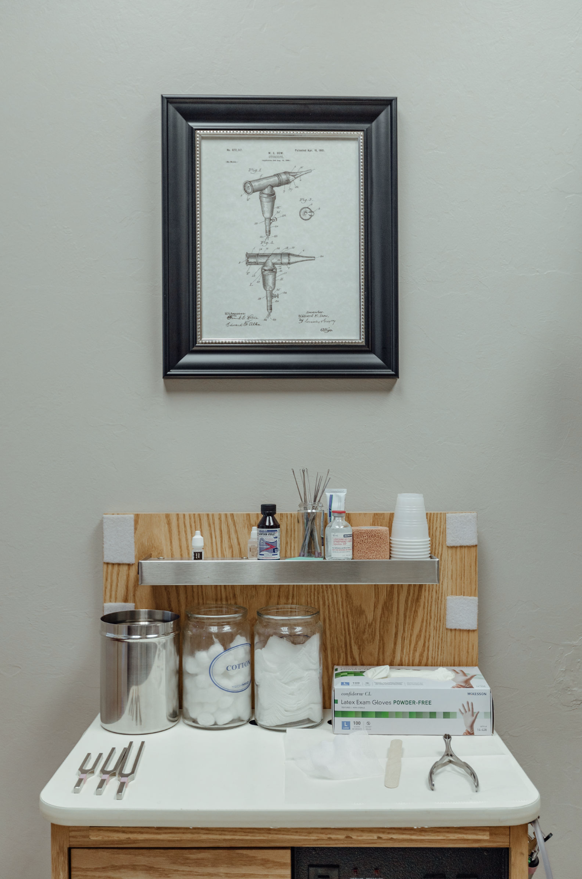 A neatly organized medical station features supplies like cotton, gauze, gloves, and tuning forks, with a framed patent diagram of a medical instrument displayed above.