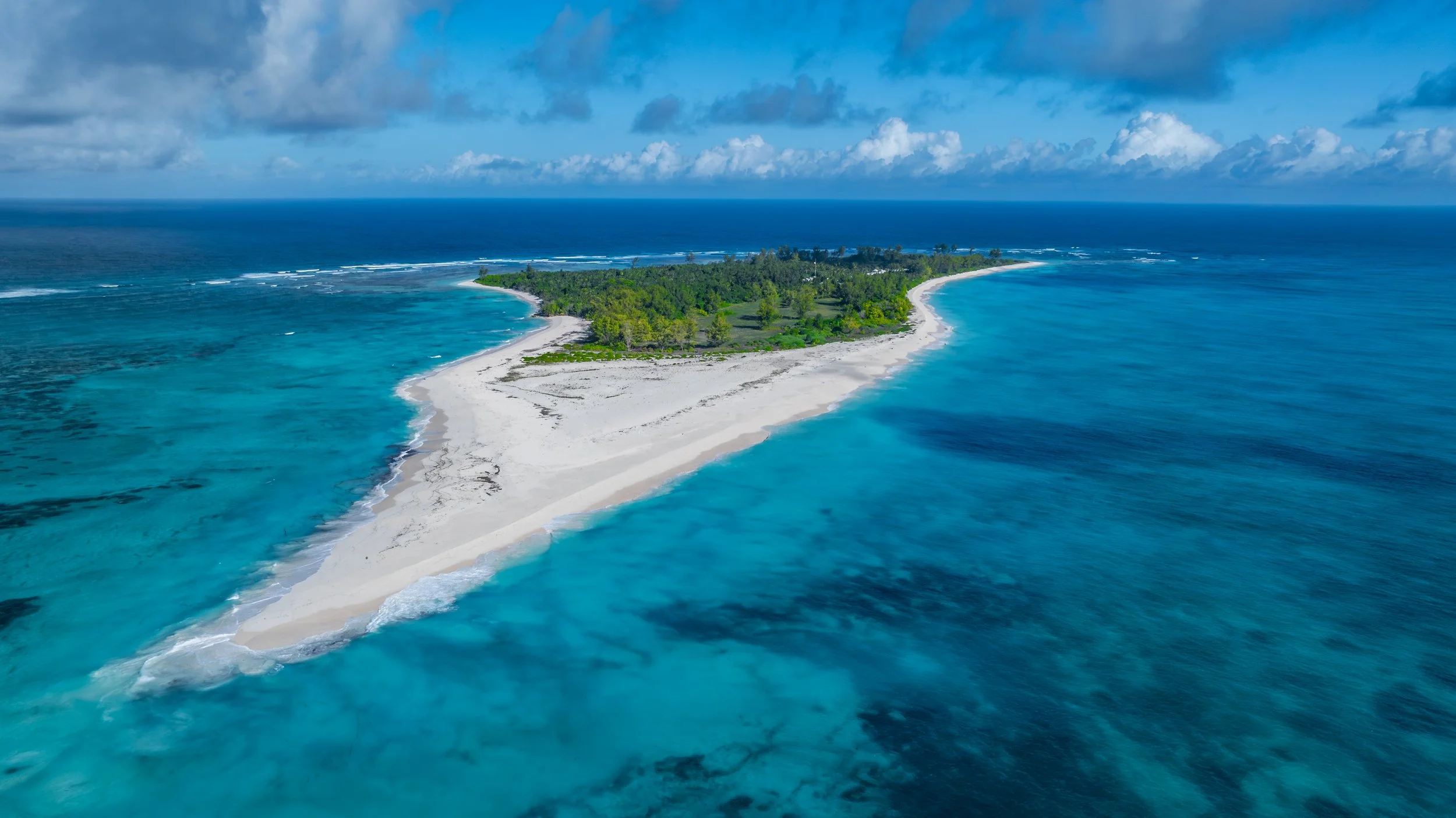Eine Insel mit weißem Strand, umgeben von türkisblauem Wasser, mit einer dichten grünen Vegetation im Zentrum, unter einem bewölkten Himmel.