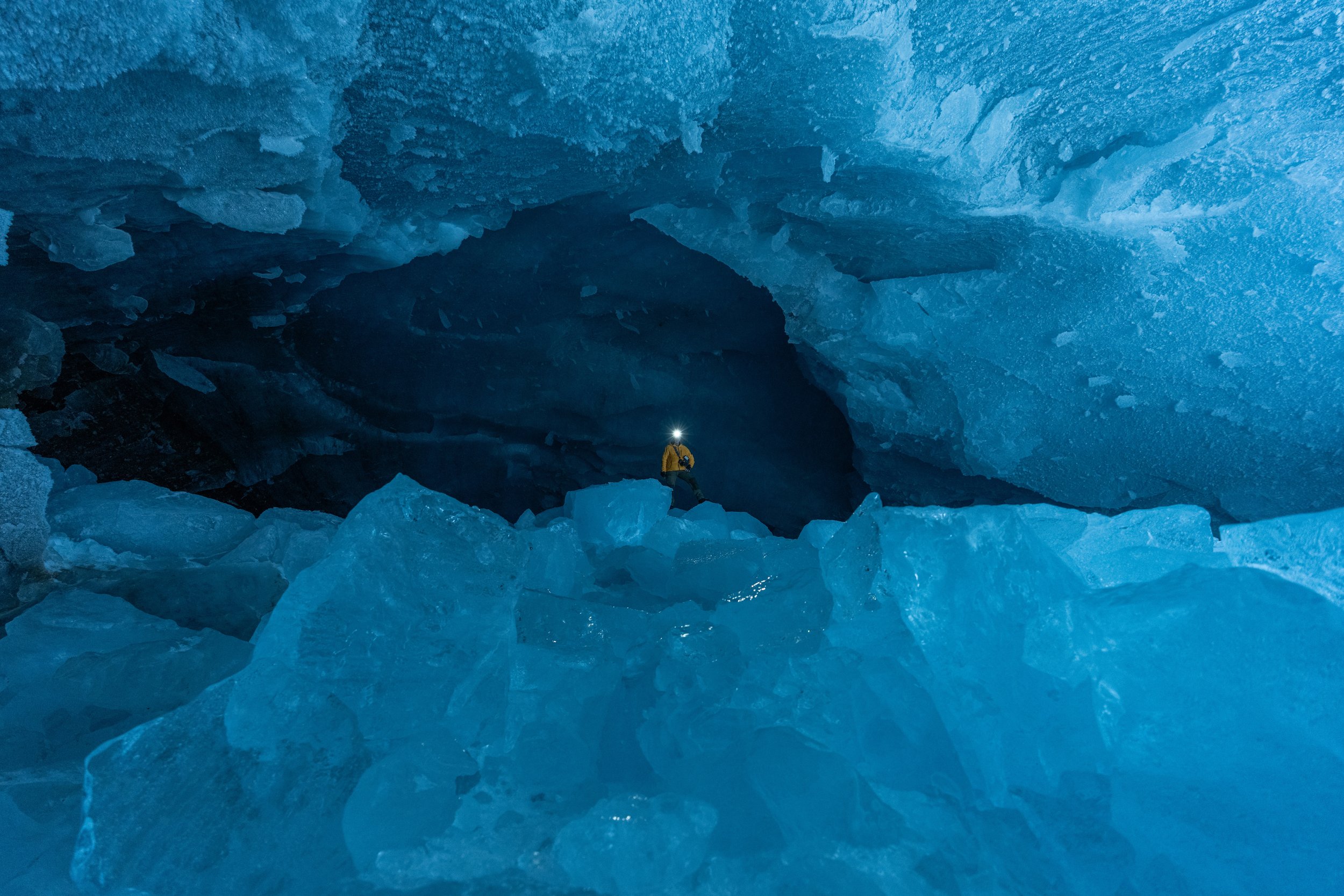 Ein Eiskletterer mit gelber Jacke und Stirnlampe in einer blauen Grotte aus Eis und Schnee.