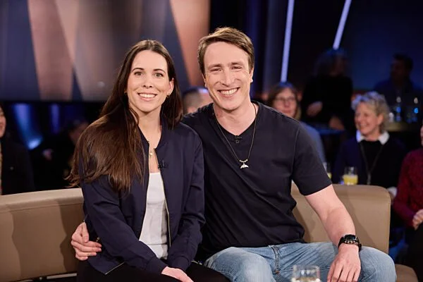 Ein Mann und eine Frau sitzen nebeneinander auf einem Sofa in einer Fernsehsendung, umgeben von einem Publikum, während sie lächeln.