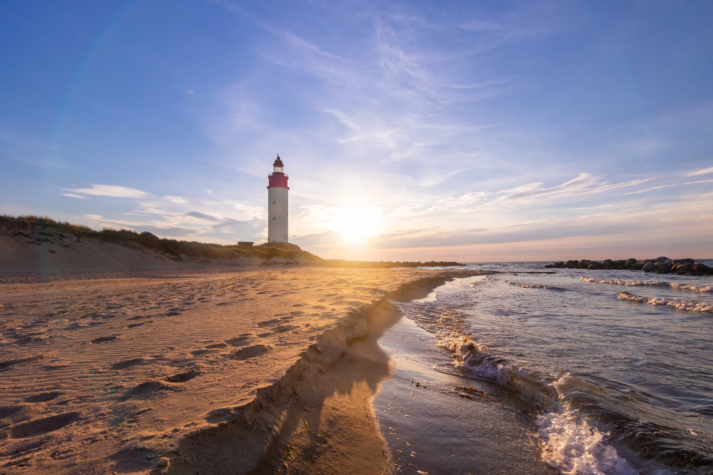 Leuchtturm am Strand bei Sonnenuntergang, mit Sand, Wasser und Himmel im Hintergrund.