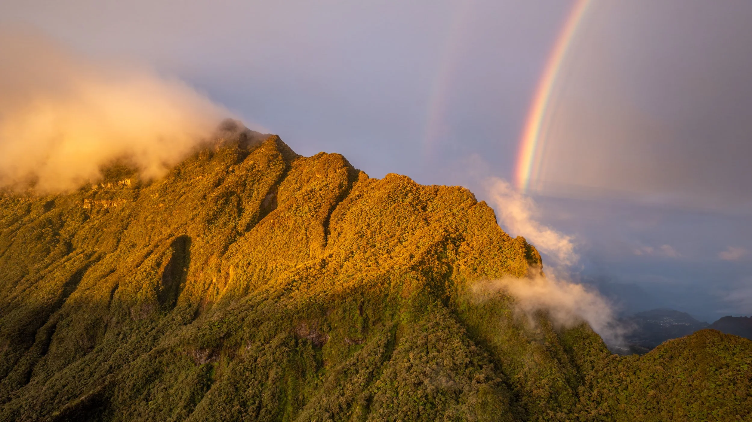 Berglandschaft mit grünem, bewachsenem Gipfel, umgeben von Wolken, bei Sonnenuntergang mit Regenbogen im Himmel.