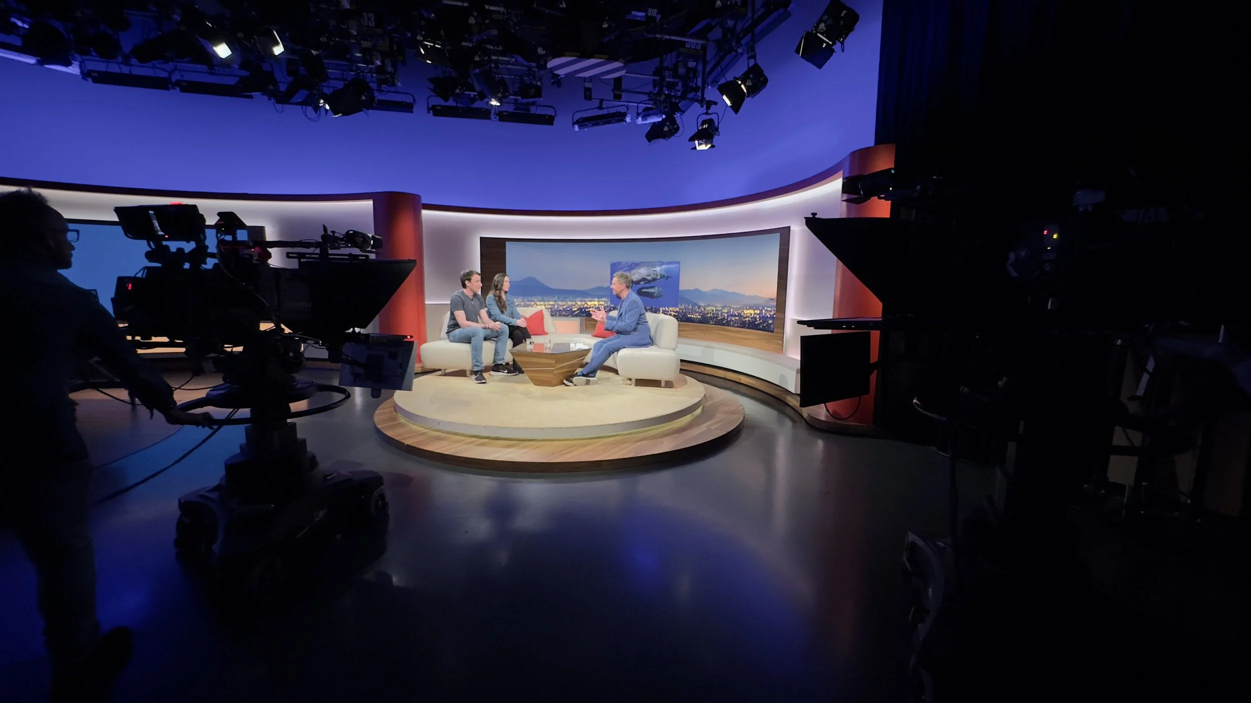 Zwei Menschen sitzen auf einem Sofa im Fernsehenstudio, mit einem Moderator. Die Kamera filmt den Bereich, während im Hintergrund eine große Bildschirmwand einen Stadtblick bei Sonnenuntergang zeigt.