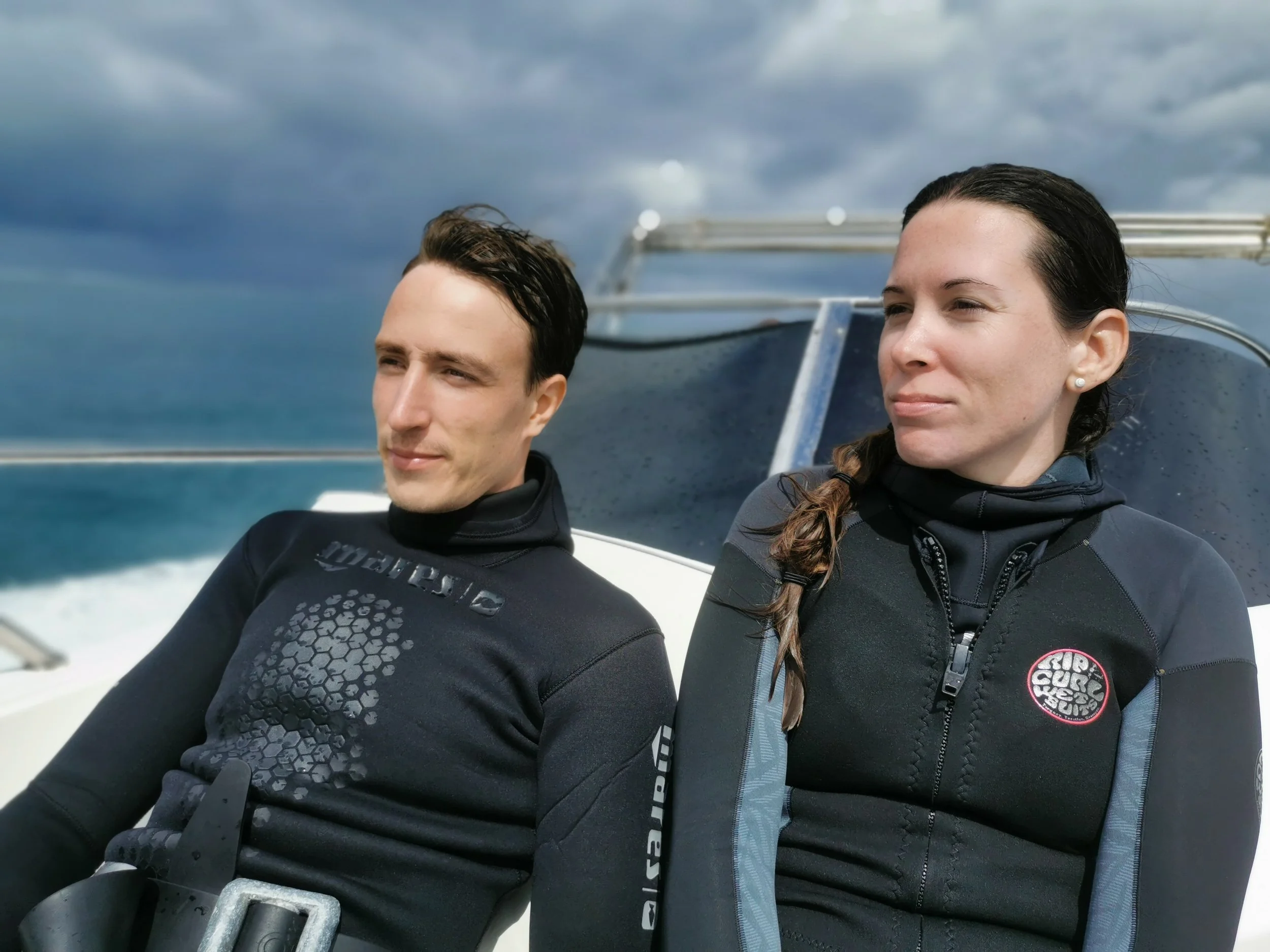 Zwei Menschen in Neoprenanzügen sitzen auf einem Boot, mit Blick auf den Himmel und das Meer.