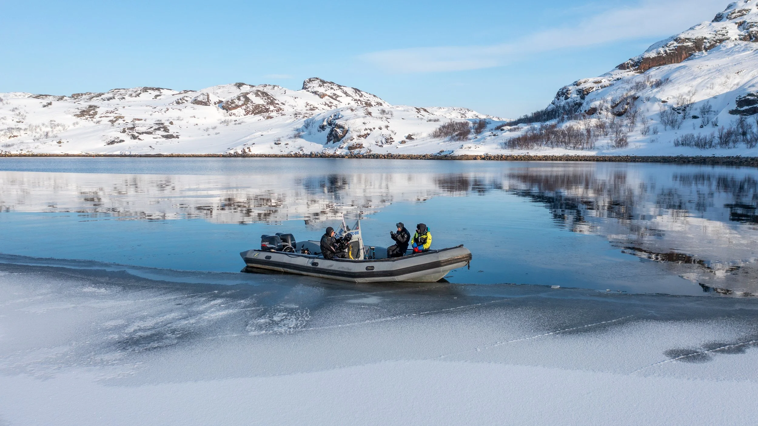 Drei Personen in einem Boot auf einem gefrorenen See, umgeben von schneebedeckten Bergen in der Ferne, bei klarem Himmel.