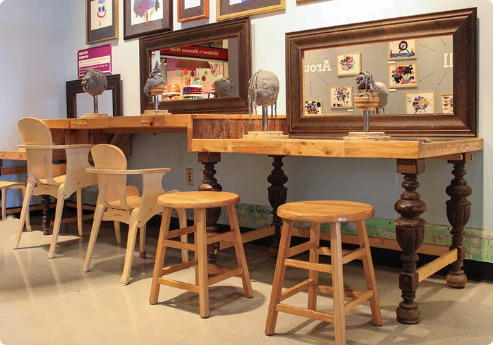 Two desks (1 child-sized and 1 grownup-sized) are topped with mounds of sculptable clay, next to sets of chairs.