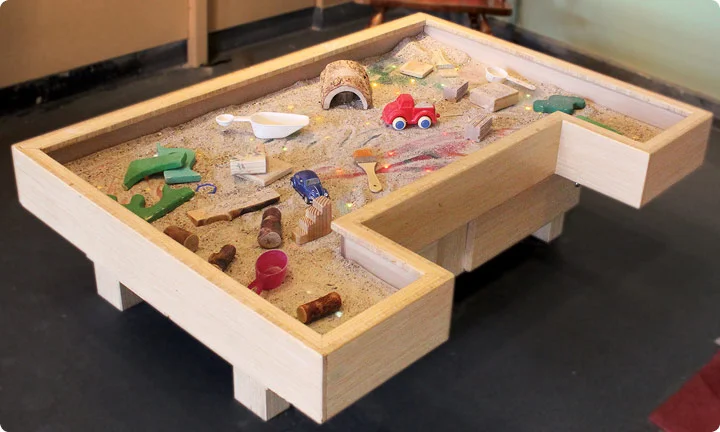 A box filled with shimmering sand, shovels, and other toys is ready for toddlers to explore.