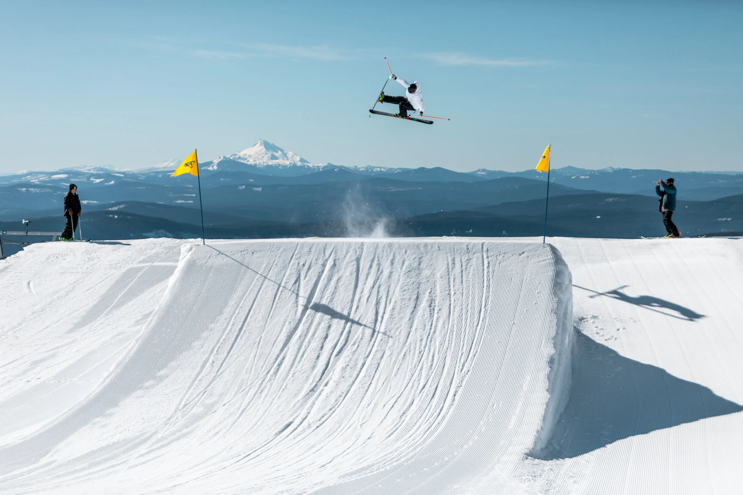 A skier performing a mid-air jump on a snow ramp in a ski park with two people watching, one taking a photo, and snow-capped mountains in the background.