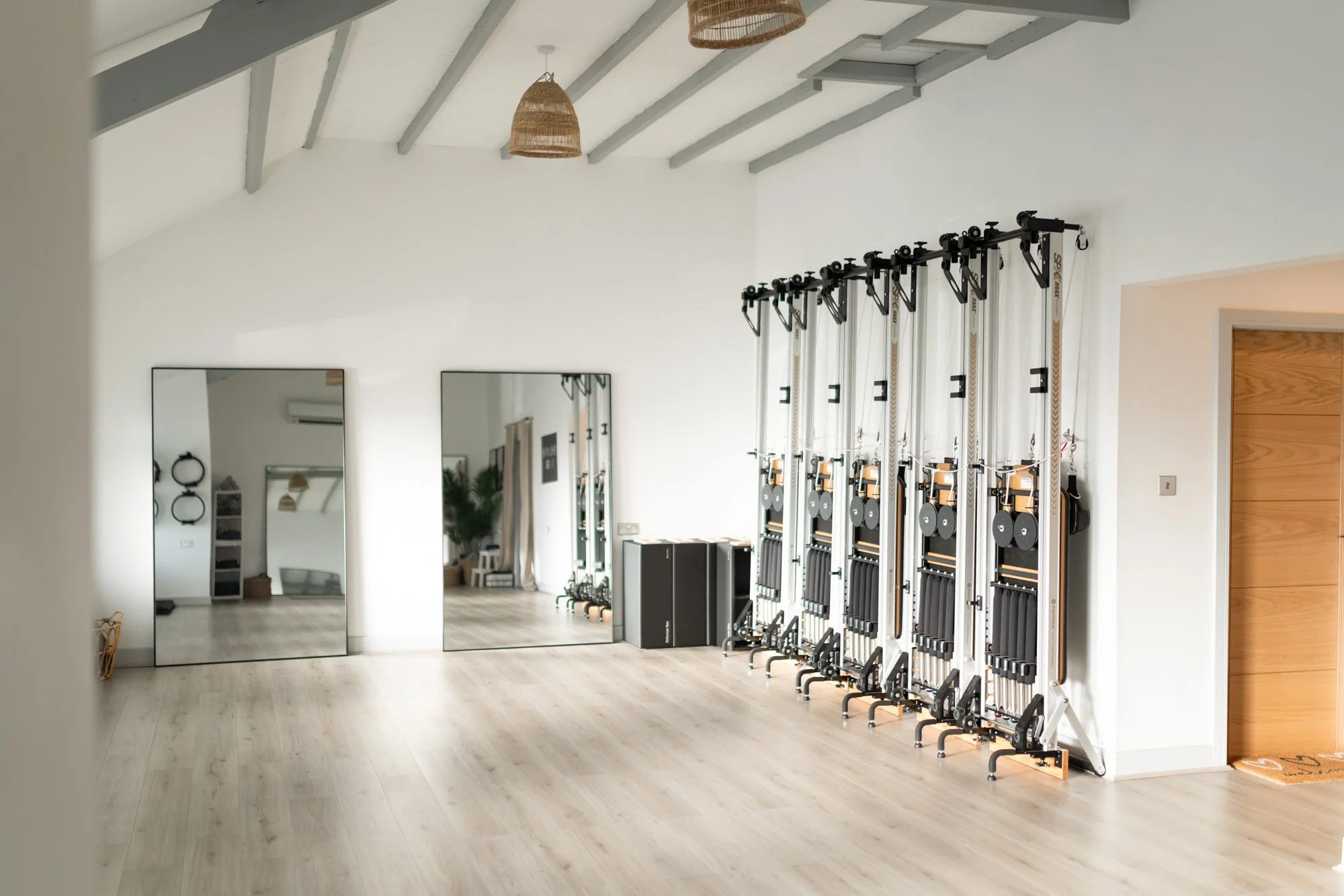 Pilates studio with wooden floors, white walls, a large mirror, and tall workout equipment with pull-up bars, weights, and resistance bands.