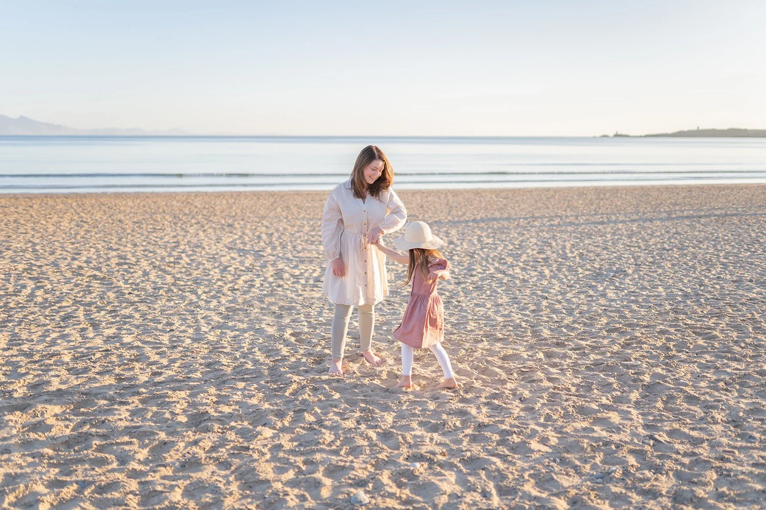 A woman and a young girl playing together on a sandy beach during sunset, with the ocean and distant mountains in the background.