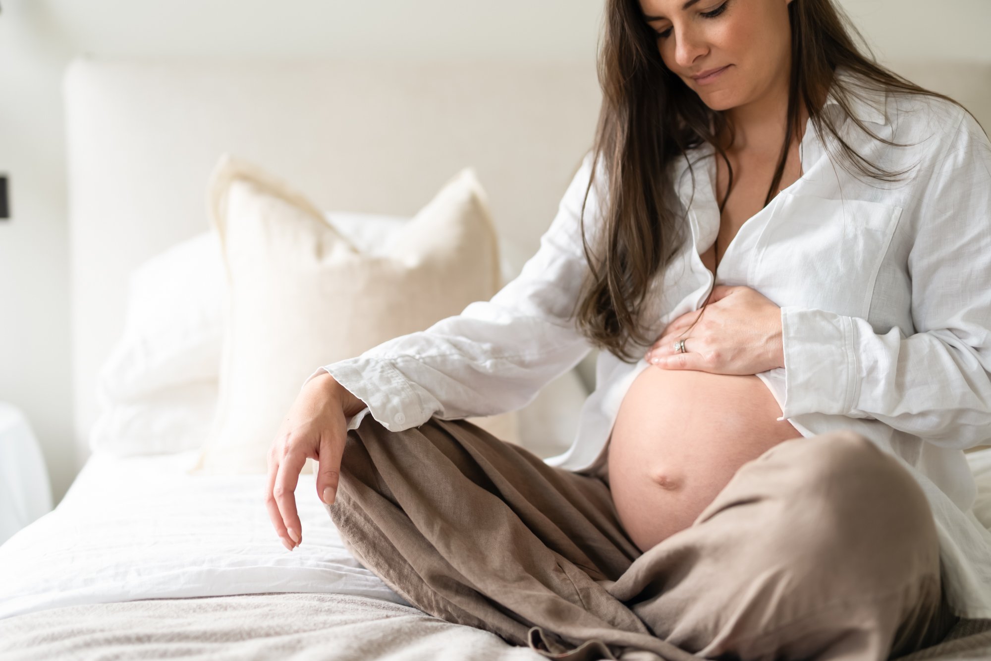 A woman sitting on a bed touching her pregnant belly, wearing an unbuttoned white shirt and brown linen trousers.
