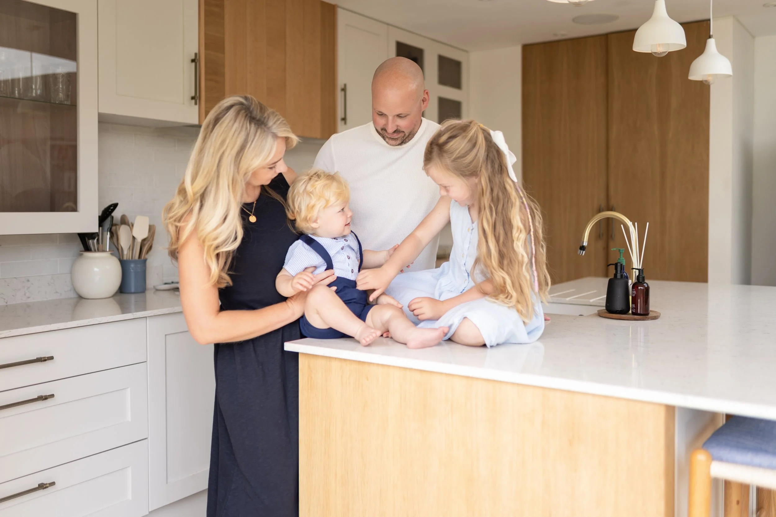 Family of five gathered in a modern kitchen, smiling and interacting, with a mother holding a toddler, an older girl reaching out, and a father standing nearby, all enjoying family time.