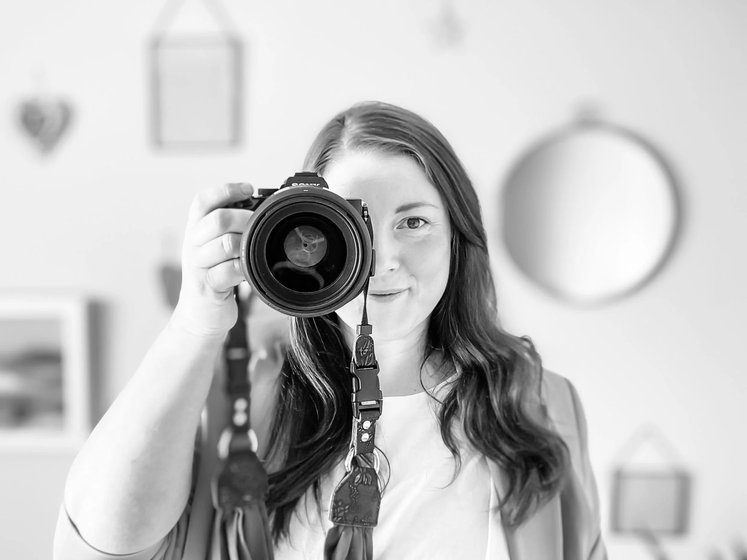 A woman with long hair takes a selfie with a camera, with small decorative objects hanging on the wall behind her.